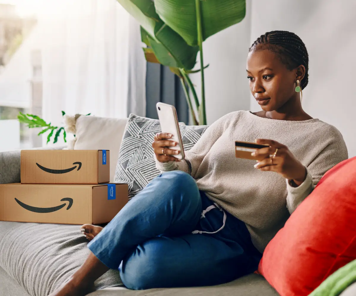 A woman sitting on a couch looks at her smartphone while holding a credit card, with Amazon Prime boxes stacked next to her.