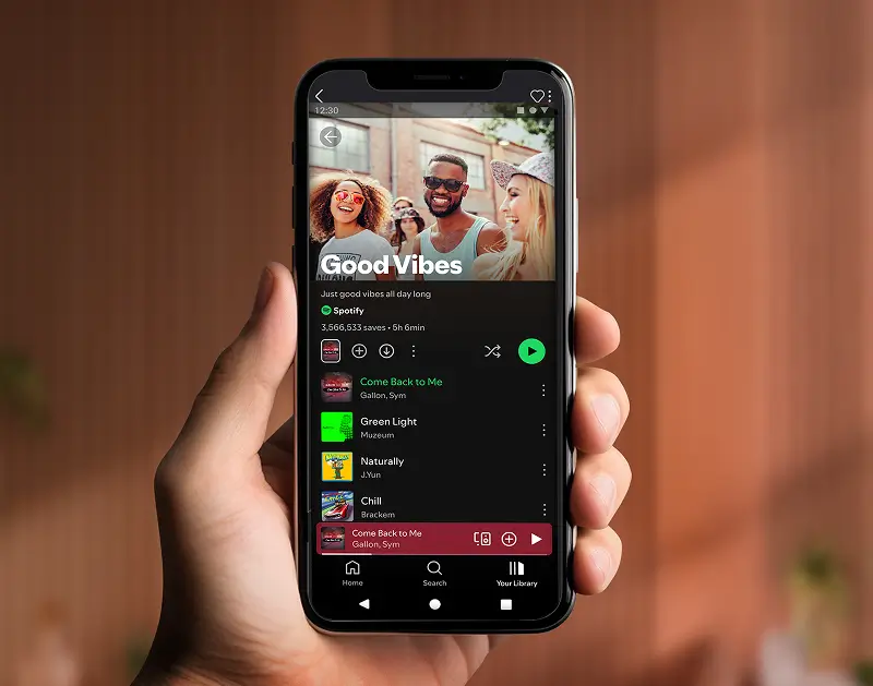 A person's hand holding a black smartphone displaying the Spotify mobile app interface. The screen shows a "Good Vibes" playlist featuring a cover image of three smiling friends outdoors. Below the playlist title, there are various song listings such as "Come Back to Me" by Gallon and Sym, and "Green Light" by Muzeum. A red playback bar is visible at the bottom of the screen, and the background behind the hand is a softly blurred indoor setting with warm lighting.