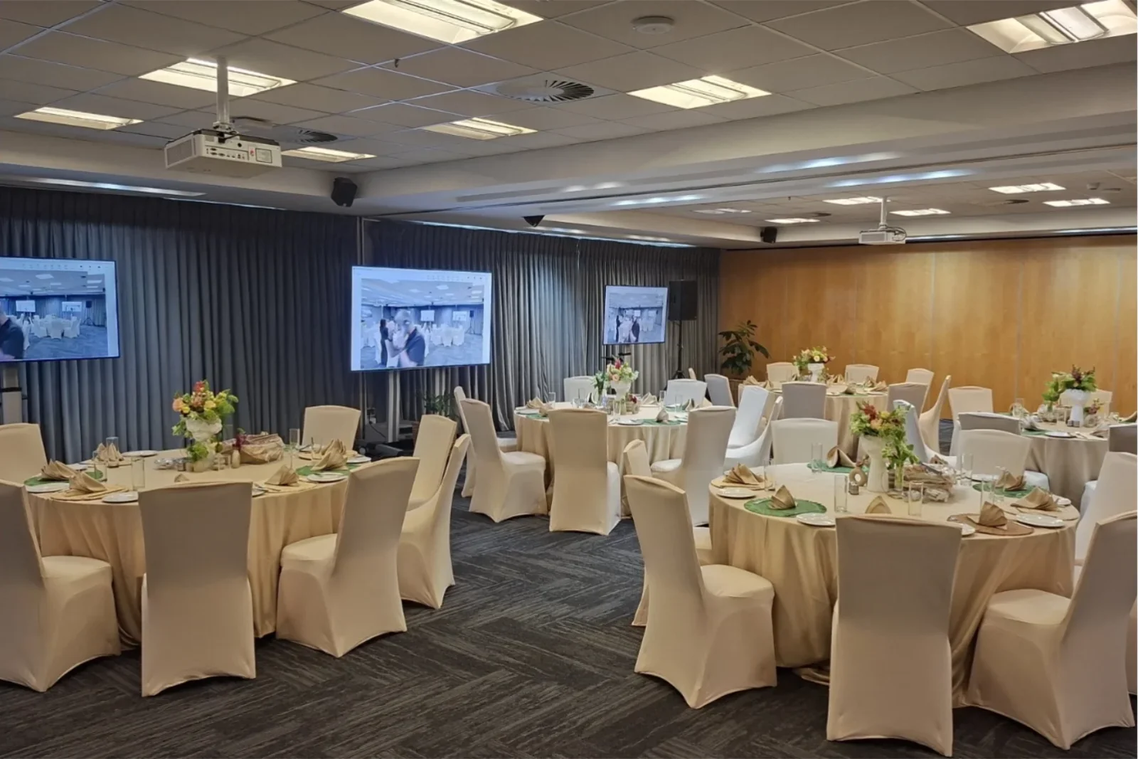 Indoor banquet hall setup with round tables, cream seat covers, and display screens